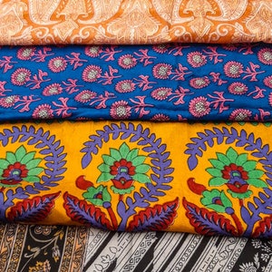 May include: A stack of five fabric swatches with different floral patterns. The top swatch is a light cream with blue flowers, the second is orange with a paisley pattern, the third is blue with pink flowers, the fourth is yellow with green and purple flowers, and the bottom is black with a gray and white striped pattern.