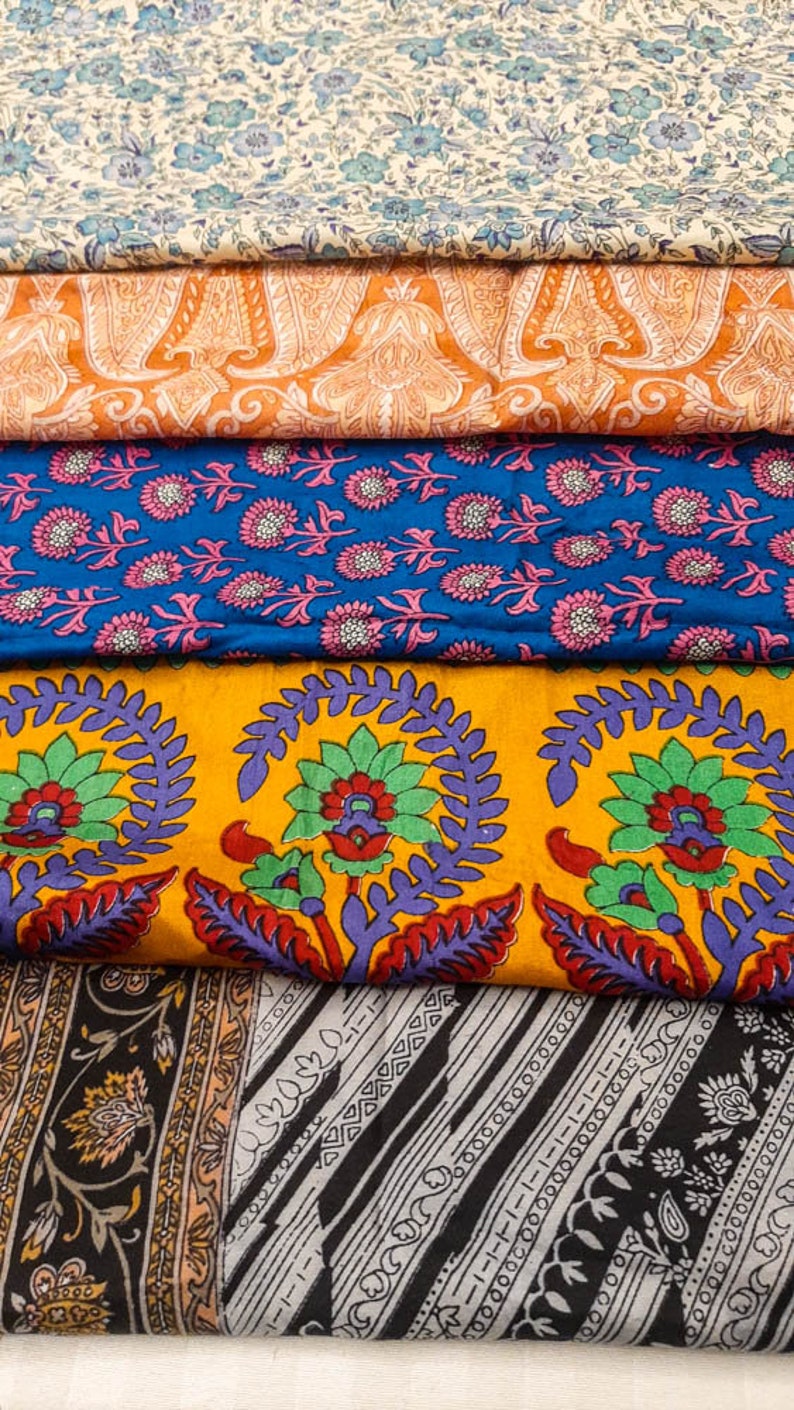May include: A stack of five fabric swatches with different floral patterns. The top swatch is a light cream with blue flowers, the second is orange with a paisley pattern, the third is blue with pink flowers, the fourth is yellow with green and purple flowers, and the bottom is black with a gray and white striped pattern.