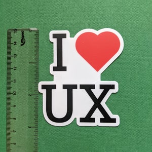 May include: A white sticker with the text "I &hearts; UX" in black letters and a red heart. A ruler with centimeter markings is on the left side of the sticker. The sticker is on a green background.