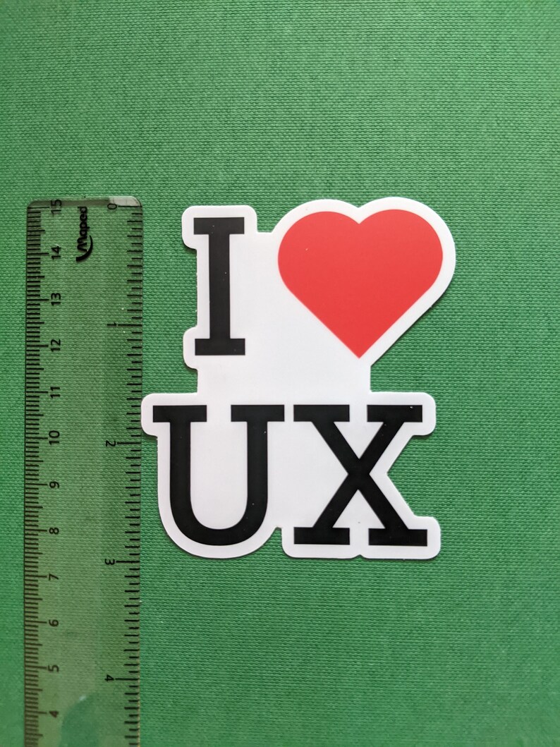 May include: A white sticker with the text "I &hearts; UX" in black letters and a red heart. A ruler with centimeter markings is on the left side of the sticker. The sticker is on a green background.