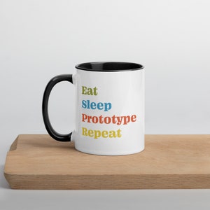 Puede incluir: Taza de cerámica blanca con borde y asa negros. La taza tiene el texto "Eat Sleep Prototype Repeat" en letras de colores.