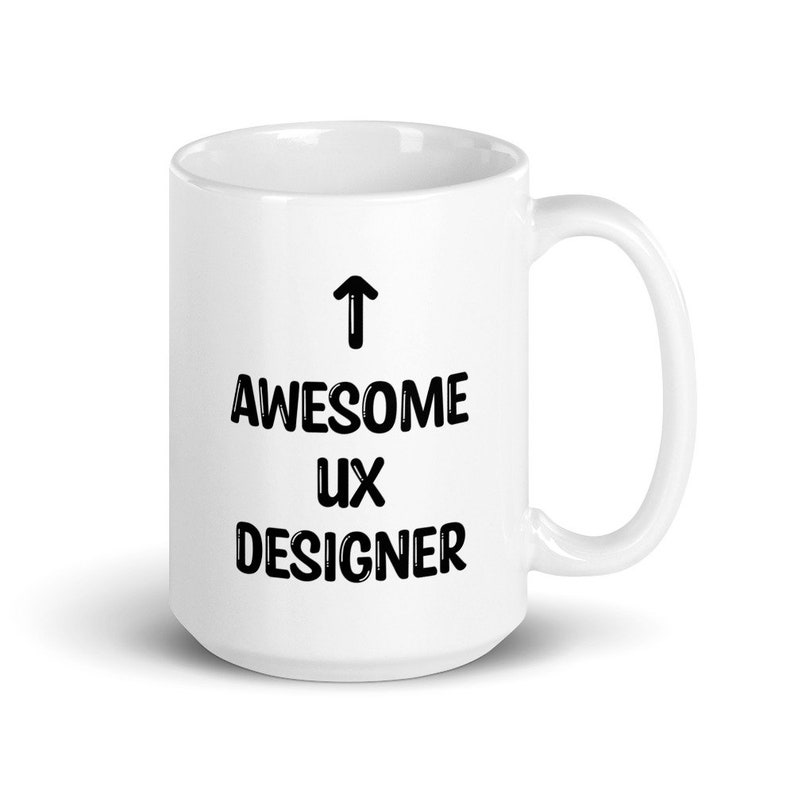 K&ouml;nnte beinhalten: Wei&szlig;e Keramik-Kaffeetasse mit schwarzem Text, der "&uarr; AWESOME UX DESIGNER" lautet.