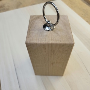 May include: A wooden doorstop with a metal ring handle. The doorstop is a light brown color and has a smooth finish.