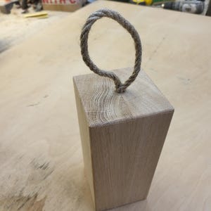 May include: A natural wood doorstop with a rope handle. The doorstop is a light brown color and has a rough texture.
