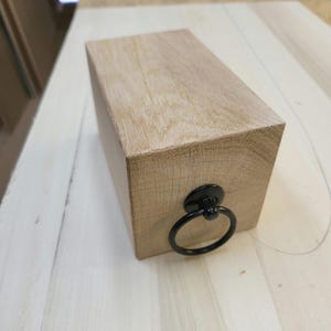 May include: A wooden box with a black metal ring handle. The box is made of light wood and has a smooth finish.