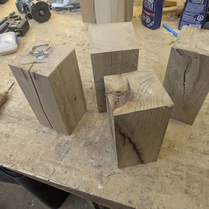 May include: Several unfinished wooden blocks of varying sizes are arranged on a workbench. One block has a metal ring attached to the top. The wood has a natural, light brown color with visible grain and some cracks.