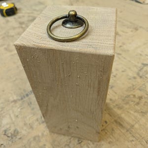 May include: A light-colored, rectangular wooden block with a brass ring pull attached to the top. The ring pull is circular and has a small, decorative base. The wood grain is visible, and the block appears to be unfinished.