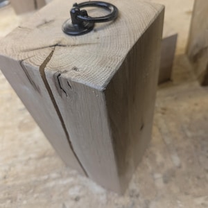 May include: A light brown, rectangular wooden block with a black metal ring pull attached to the top. The wood grain is visible, and a crack runs down one side. The block is likely a furniture component or decorative item.