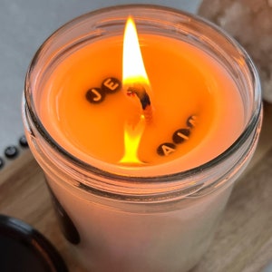 May include: A glass jar candle with a lit wick and the words "JE AIM" spelled out in black letters on the surface of the wax.