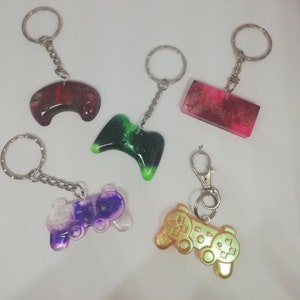 May include: Five different resin keychains shaped like video game controllers. The keychains are in various colours, including green, purple, gold, and pink.