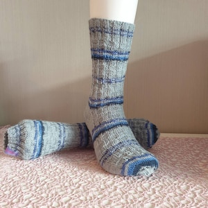 Könnte beinhalten: Ein Paar graue und blaue gestreifte Socken mit einem strukturierten Strickmuster. Die Socken sind gefaltet und auf einer rosa gehäkelten Oberfläche ausgestellt.