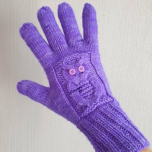 Könnte beinhalten: Lilafarbene Strickhandschuhe mit einem Eulenmotiv auf dem Handrücken. Die Eule hat zwei rosa Knöpfe für die Augen.