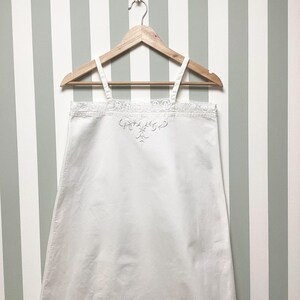 May include: A white sleeveless garment with thin shoulder straps, featuring a delicate lace trim along the upper edge and a subtle white embroidered floral motif centred just below the neckline, all displayed on a wooden clothes hanger.