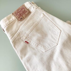 Pode incluir: Close-up de jeans Levi's 501 bege. Os jeans têm um design clássico de cinco bolsos com uma etiqueta vermelha Levi's no bolso de trás. A etiqueta Levi Strauss & Co. é visível na cintura. Os jeans são feitos de um material de ganga resistente.