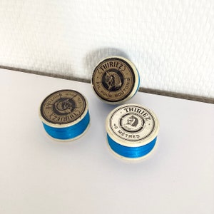 Puede incluir: Tres carretes de hilo azul con etiquetas blancas. Las etiquetas tienen el texto "Thiriez" y "Fil pour boutons". Un carrete tiene el texto "40 metros".