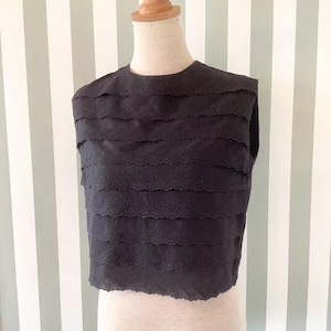 Puede incluir: Un top negro sin mangas con un diseño en capas. El top, exhibido en un maniquí, tiene un cuello redondo y bordes festoneados. La tela tiene un sutil patrón de puntos.