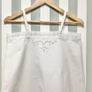 Op de afbeelding: Een wit mouwloos hemdje met dunne schouderbandjes en een fijne witte kanten rand langs de rechte halslijn. Het midden van de borst is versierd met een subtiel wit bloemenborduurpatroon in een symmetrisch ontwerp.