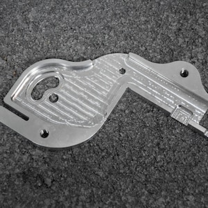 May include: A silver metal part with a textured surface and several holes. The part is shaped like a curved L and has a small rectangular cutout on the right side.