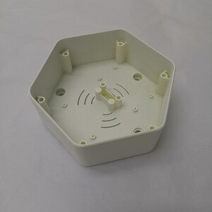 以下が含まれることがあります： 中央に小さな白いプラスチック製の部品が付いた、白い六角形のプラスチック製の箱。箱の縁には小さな穴がいくつかあり、中央には波状の線模様があります。