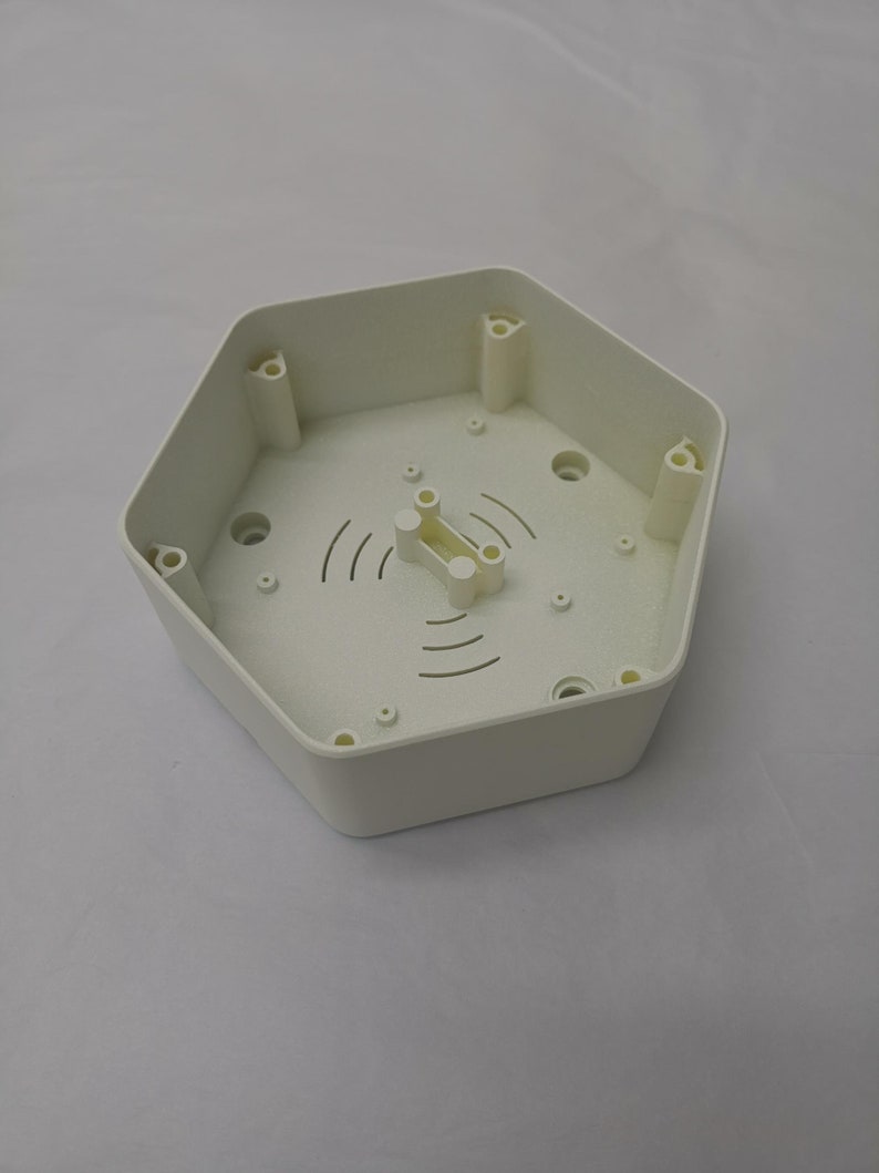以下が含まれることがあります： 中央に小さな白いプラスチック製の部品が付いた、白い六角形のプラスチック製の箱。箱の縁には小さな穴がいくつかあり、中央には波状の線模様があります。