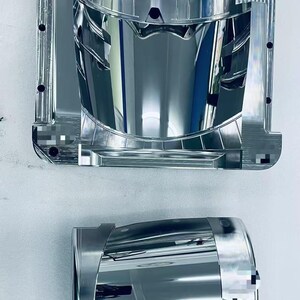 Peut inclure: Deux pièces automobiles métalliques argentées et brillantes sont présentées sur fond blanc. La pièce supérieure est un grand moule rectangulaire avec un intérieur incurvé. La pièce inférieure est un composant cylindrique, également argenté et réfléchissant.