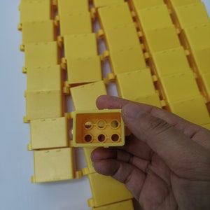 May include: A close-up of a yellow plastic block with six holes in the center. The block is being held in a hand. There are many other similar blocks in the background.