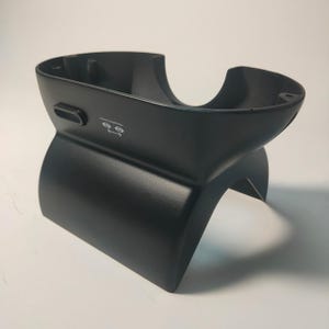 May include: A matte black plastic headset stand with a curved, open-top cradle design. The front features a small, horizontal rectangular button and a white icon depicting two eyes with a double-headed arrow between them, indicating an adjustment.