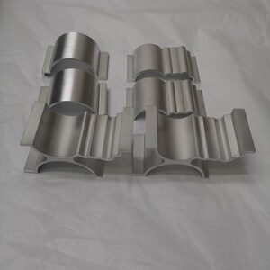 May include: Five silver metal pipe clamps with a decorative design. The clamps are made of a smooth, polished metal and have a slightly curved shape.