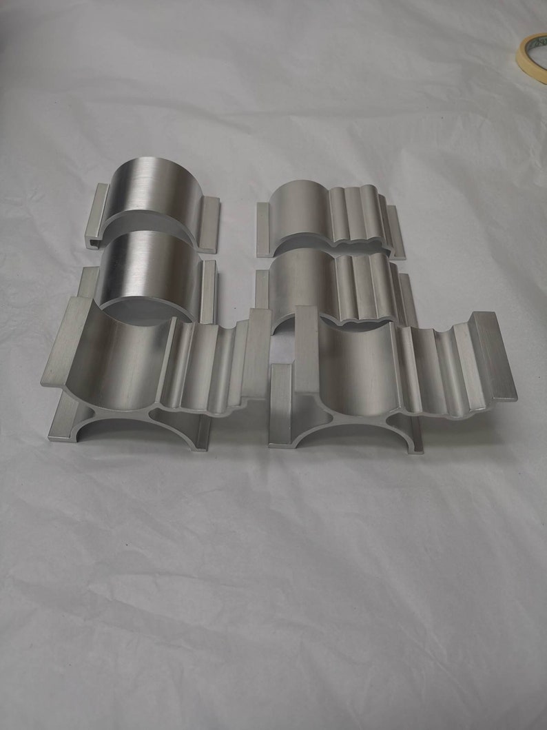 May include: Five silver metal pipe clamps with a decorative design. The clamps are made of a smooth, polished metal and have a slightly curved shape.