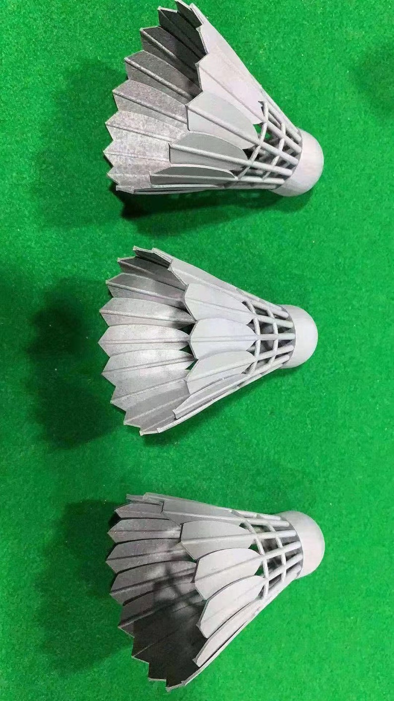Puede incluir: Tres volantes de b&aacute;dminton plateados con plumas blancas y una red de malla negra. Los volantes est&aacute;n dispuestos en fila sobre una superficie verde.