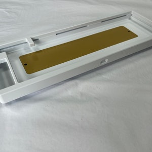 May include: A rectangular white mechanical keyboard case featuring a smooth matte finish and a centred, elongated gold-coloured metal plate. The interior structure includes precise cutouts for switches and stabilisers, showcasing a clean, hollow frame design.