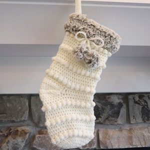 May include: A cream-colored, handmade Christmas stocking with a textured, bobble stitch design. The top is trimmed with faux fur and adorned with a bow and small pom-poms. The stocking is hanging from a white surface.