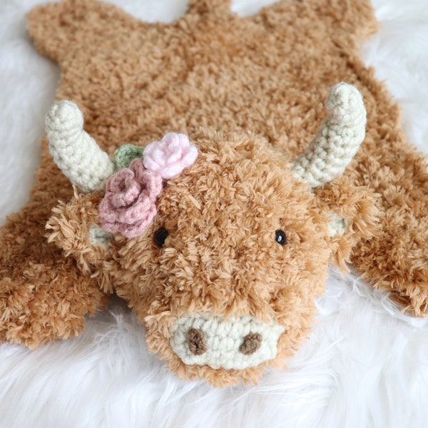 May include: A plush, brown Highland cow rug with a crocheted face, horns, and a floral crown. The rug has a shaggy texture and is designed to resemble the animal's shape. The horns are off-white, and the flowers are pink and green.