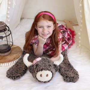 May include: A young girl with red hair is lying on a brown and white crocheted cow rug. She is wearing a pink headband and a plaid dress. The rug has a pink flower on its head.