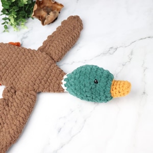 May include: A brown crocheted duck plush toy with a green head and orange beak. The duck is lying on its back with its wings spread out.