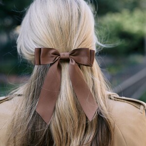 May include: A brown satin bow hair accessory is secured in blonde hair. The bow is a rich, warm brown color and is tied in a classic bow shape. The hair accessory adds a touch of elegance.