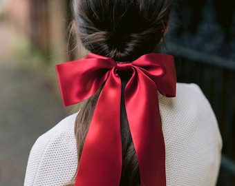May include: A large, vibrant red satin bow is tied in the hair. The bow is the focal point, with long, flowing tails. The person is wearing a white sweater. The background is blurred, suggesting an outdoor setting.