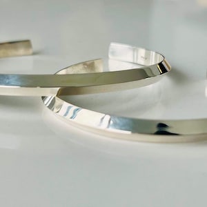 Peut inclure: Deux bracelets en argent avec un design plat, large et légèrement angulaire. Les bracelets sont empilés l'un sur l'autre.