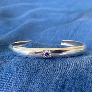 Peut inclure: Un bracelet manchette argenté poli avec une petite pierre précieuse ronde bleu foncé au centre. Le bracelet a un design lisse et incurvé. Il est présenté sur une surface en denim bleu.