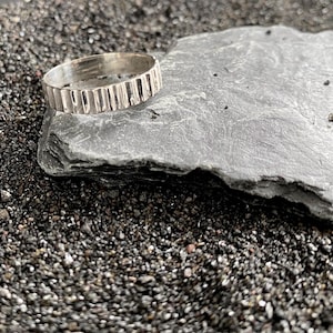 Peut inclure: Une bague en argent avec un motif texturé de lignes verticales. La bague est posée sur une pierre grise, elle-même sur un lit de petits galets sombres. La surface de la bague reflète la lumière, soulignant son motif détaillé. L'esthétique générale est minimaliste et moderne.