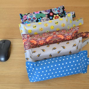 May include: Five fabric pouches with different patterns, including skulls, smiley faces, flowers, bees, and stars. The pouches are stacked on top of each other and are arranged in a row. A black computer mouse is in the lower left corner of the image.