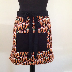 May include: A black apron with a festive print of carrots wearing Santa hats. The apron has two pockets and a black tie.