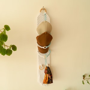 May include: White macrame wall hanging with a blue and white beaded rope, two brown baseball caps, and a colourful fabric hanging from the bottom.