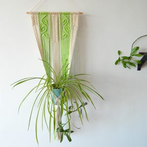 Peut inclure: Un porte-plante en macramé avec un schéma de couleurs vert et crème. Le porte-plante est fait de cordon de coton et a une baguette en bois en haut. Une plante verte est suspendue dans le pot.
