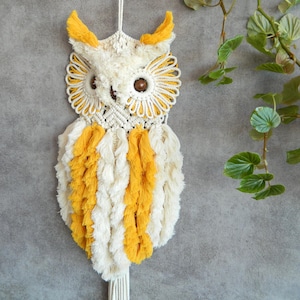 May include: A handmade macrame owl wall hanging. The owl features a cream-colored body with yellow accents on the ears and chest. The eyes are brown, and the design incorporates intricate knotting patterns. A decorative piece for home decor.