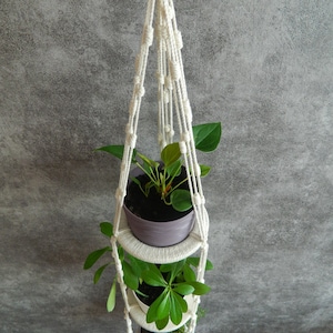 Pode incluir: Um suporte de plantas de macramé branco com três níveis, cada um com uma planta em vaso. As plantas são verdes e frondosas, e o suporte está pendurado no teto.