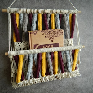 May include: A macrame wall hanging with a book shelf. The macrame is made with white, grey, yellow, and burgundy yarn. The shelf is made of wood and has a white bead on each end.