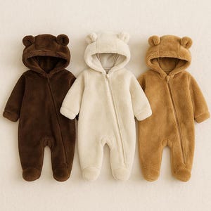 May include: Three baby onesies in shades of brown, cream, and tan. Each has a hood with bear ears and a full-length zip. The soft, fluffy material covers the arms and legs, creating a cosy outfit for infants.