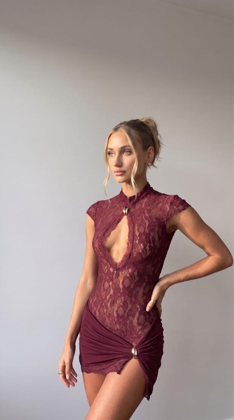 Puede incluir: Un vestido corto de encaje color burdeos con cuello alto y abertura en forma de l&aacute;grima, mangas cortas y una falda fruncida con una abertura lateral asegurada por un detalle de anillo met&aacute;lico dorado en la cintura.