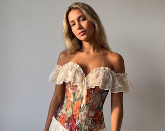 Puede incluir: Un top tipo corsé con un estampado floral vibrante en tonos naranja, rojo y verde, acentuado por un escote con volantes de color crema que deja los hombros al descubierto y una falda a juego con cintura fruncida y tela suave y fluida.